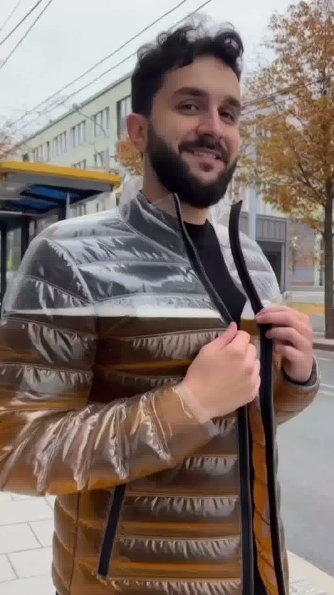Model standing in urban setting wearing the transparent Beer Puff Jacket, with visible beverage-filled chambers in a puff design, golden liquid and foamy sections clearly seen through the glossy outer shell. The high collar, full-front zip and drink-dispensing valve at chest level are visible. Sunlight reflects off the material showing the clear structure. The wearer is dressed casually underneath, letting the jacket dominate the look. The image captures the jacket in motion, drink inside settling slightly,
