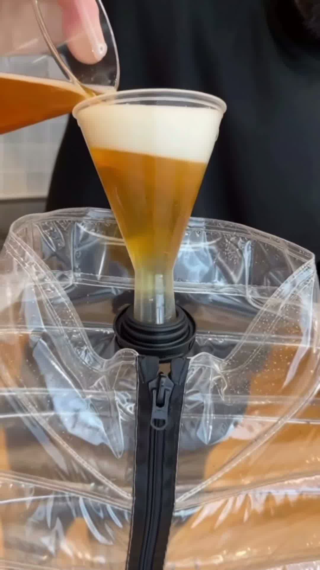 Model standing in urban setting wearing the transparent Beer Puff Jacket, with visible beverage-filled chambers in a puff design, golden liquid and foamy sections clearly seen through the glossy outer shell. The high collar, full-front zip and drink-dispensing valve at chest level are visible. Sunlight reflects off the material showing the clear structure. The wearer is dressed casually underneath, letting the jacket dominate the look. The image captures the jacket in motion, drink inside settling slightly,