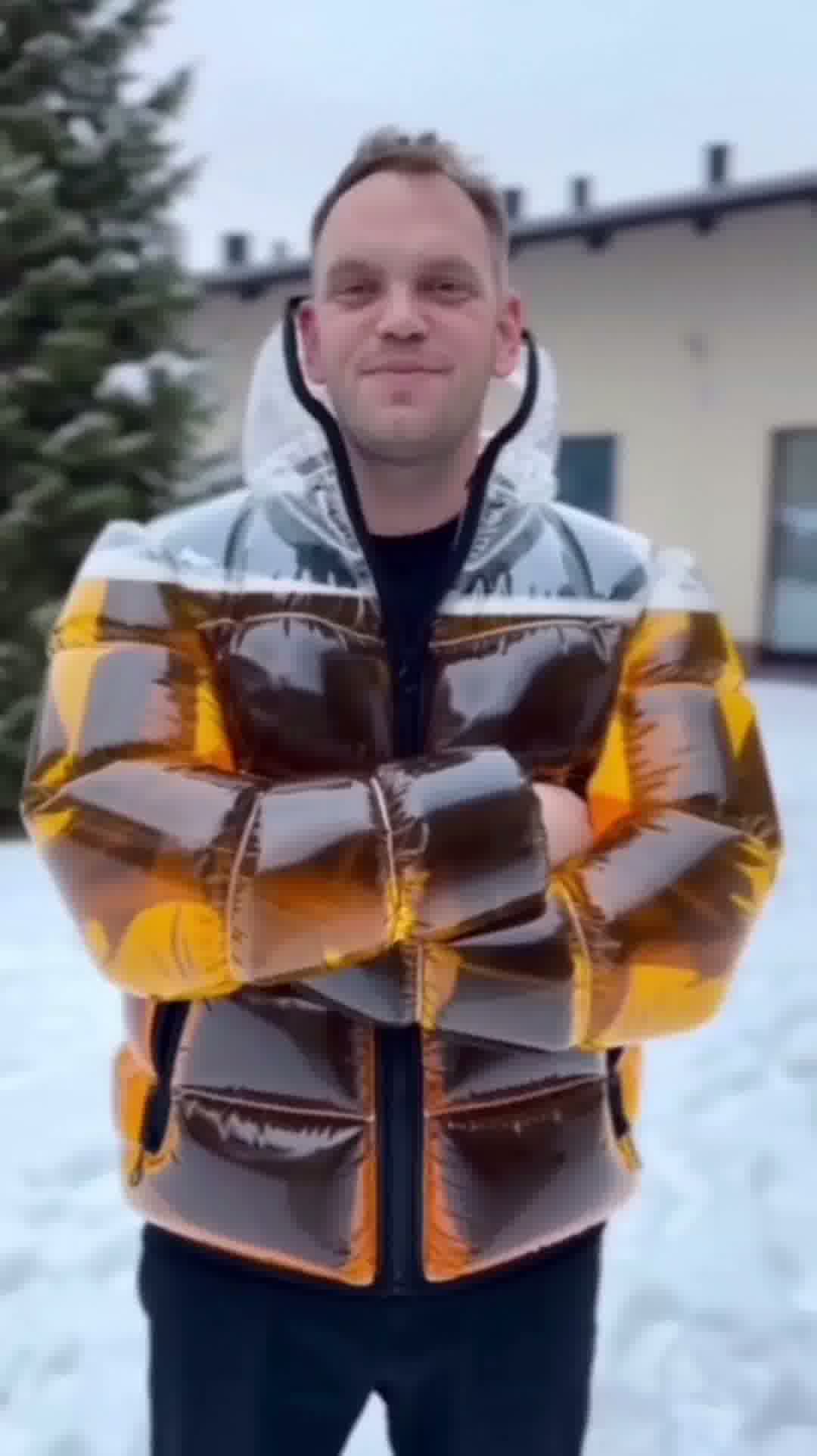 Model standing in urban setting wearing the transparent Beer Puff Jacket, with visible beverage-filled chambers in a puff design, golden liquid and foamy sections clearly seen through the glossy outer shell. The high collar, full-front zip and drink-dispensing valve at chest level are visible. Sunlight reflects off the material showing the clear structure. The wearer is dressed casually underneath, letting the jacket dominate the look. The image captures the jacket in motion, drink inside settling slightly,