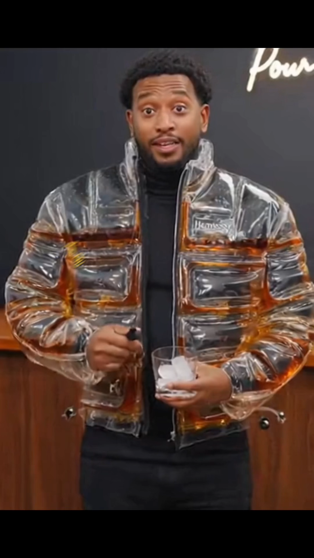 Model standing in urban setting wearing the transparent Beer Puff Jacket, with visible beverage-filled chambers in a puff design, golden liquid and foamy sections clearly seen through the glossy outer shell. The high collar, full-front zip and drink-dispensing valve at chest level are visible. Sunlight reflects off the material showing the clear structure. The wearer is dressed casually underneath, letting the jacket dominate the look. The image captures the jacket in motion, drink inside settling slightly,