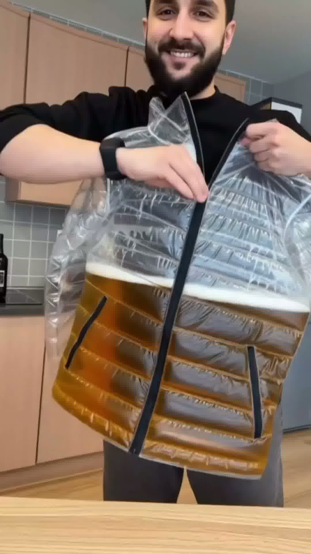 Model standing in urban setting wearing the transparent Beer Puff Jacket, with visible beverage-filled chambers in a puff design, golden liquid and foamy sections clearly seen through the glossy outer shell. The high collar, full-front zip and drink-dispensing valve at chest level are visible. Sunlight reflects off the material showing the clear structure. The wearer is dressed casually underneath, letting the jacket dominate the look. The image captures the jacket in motion, drink inside settling slightly,