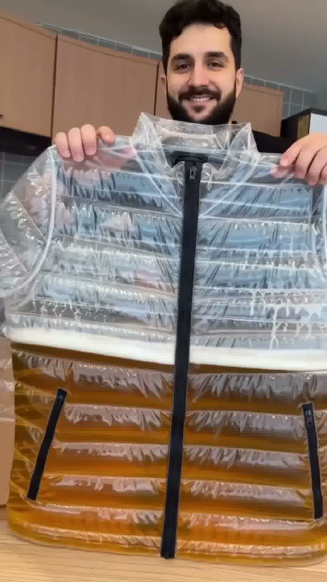 Model standing in urban setting wearing the transparent Beer Puff Jacket, with visible beverage-filled chambers in a puff design, golden liquid and foamy sections clearly seen through the glossy outer shell. The high collar, full-front zip and drink-dispensing valve at chest level are visible. Sunlight reflects off the material showing the clear structure. The wearer is dressed casually underneath, letting the jacket dominate the look. The image captures the jacket in motion, drink inside settling slightly,