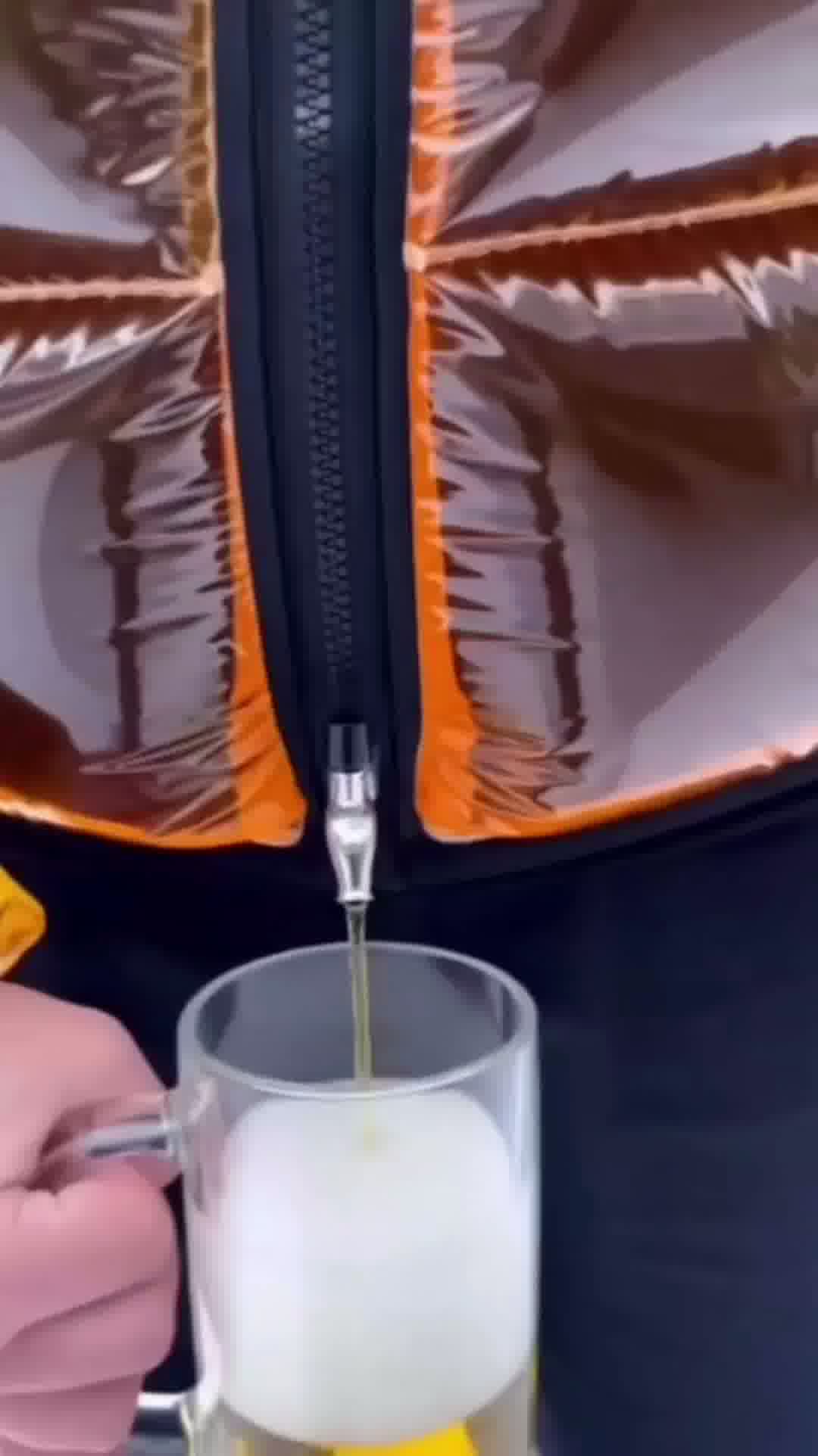 Model standing in urban setting wearing the transparent Beer Puff Jacket, with visible beverage-filled chambers in a puff design, golden liquid and foamy sections clearly seen through the glossy outer shell. The high collar, full-front zip and drink-dispensing valve at chest level are visible. Sunlight reflects off the material showing the clear structure. The wearer is dressed casually underneath, letting the jacket dominate the look. The image captures the jacket in motion, drink inside settling slightly,