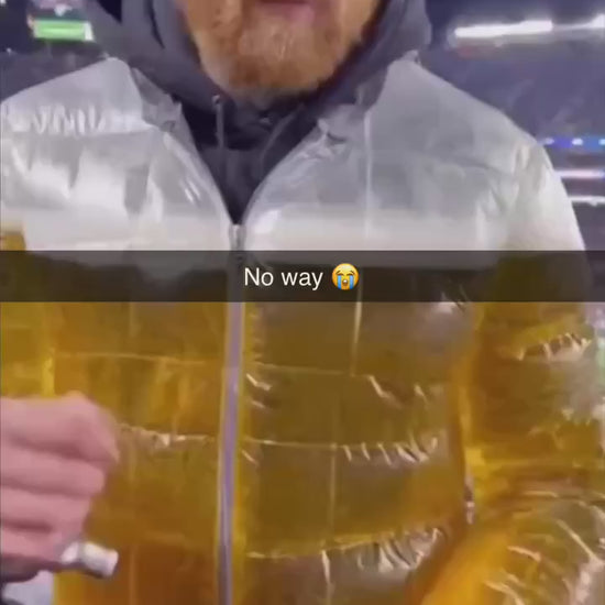 Model standing in urban setting wearing the transparent Beer Puff Jacket, with visible beverage-filled chambers in a puff design, golden liquid and foamy sections clearly seen through the glossy outer shell. The high collar, full-front zip and drink-dispensing valve at chest level are visible. Sunlight reflects off the material showing the clear structure. The wearer is dressed casually underneath, letting the jacket dominate the look. The image captures the jacket in motion, drink inside settling slightly,