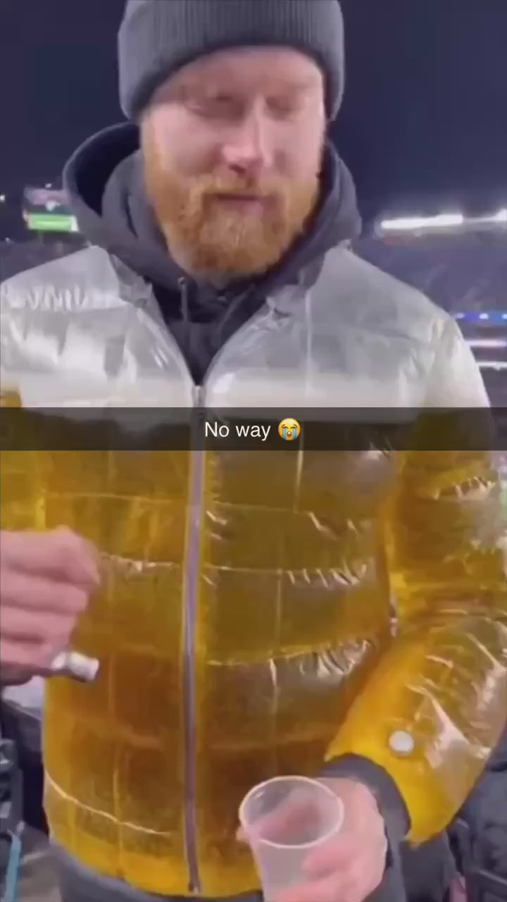 Model standing in urban setting wearing the transparent Beer Puff Jacket, with visible beverage-filled chambers in a puff design, golden liquid and foamy sections clearly seen through the glossy outer shell. The high collar, full-front zip and drink-dispensing valve at chest level are visible. Sunlight reflects off the material showing the clear structure. The wearer is dressed casually underneath, letting the jacket dominate the look. The image captures the jacket in motion, drink inside settling slightly,